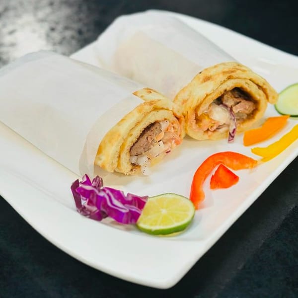 Two Afghan wraps, partially unwrapped, filled with meat and vegetables, garnished with lime and peppers.